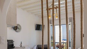 A modern living room with a kitchen area, featuring a large arched doorway, a hanging rope decoration, and a view of the outdoors. - Secret Garden Apartments (Mykonos)