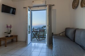 A room with a grey sofa, a wooden bench, a potted plant, and a view of the sea through an open door.