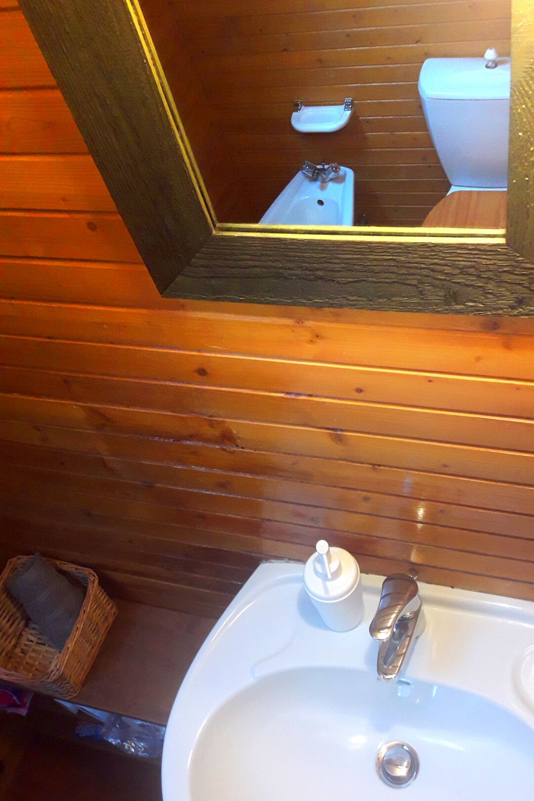 Un baño con paneles de madera, un lavabo blanco con dispensador de jabón y un espejo que refleja un inodoro y un estante pequeño.
