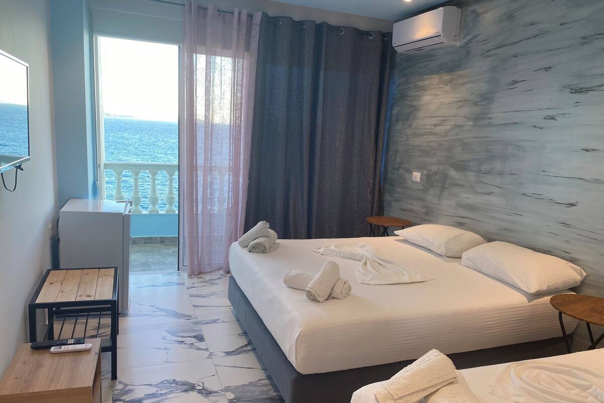 A hotel room with a bed, two bedside tables, a desk with a chair, and a view of the sea through a balcony door.