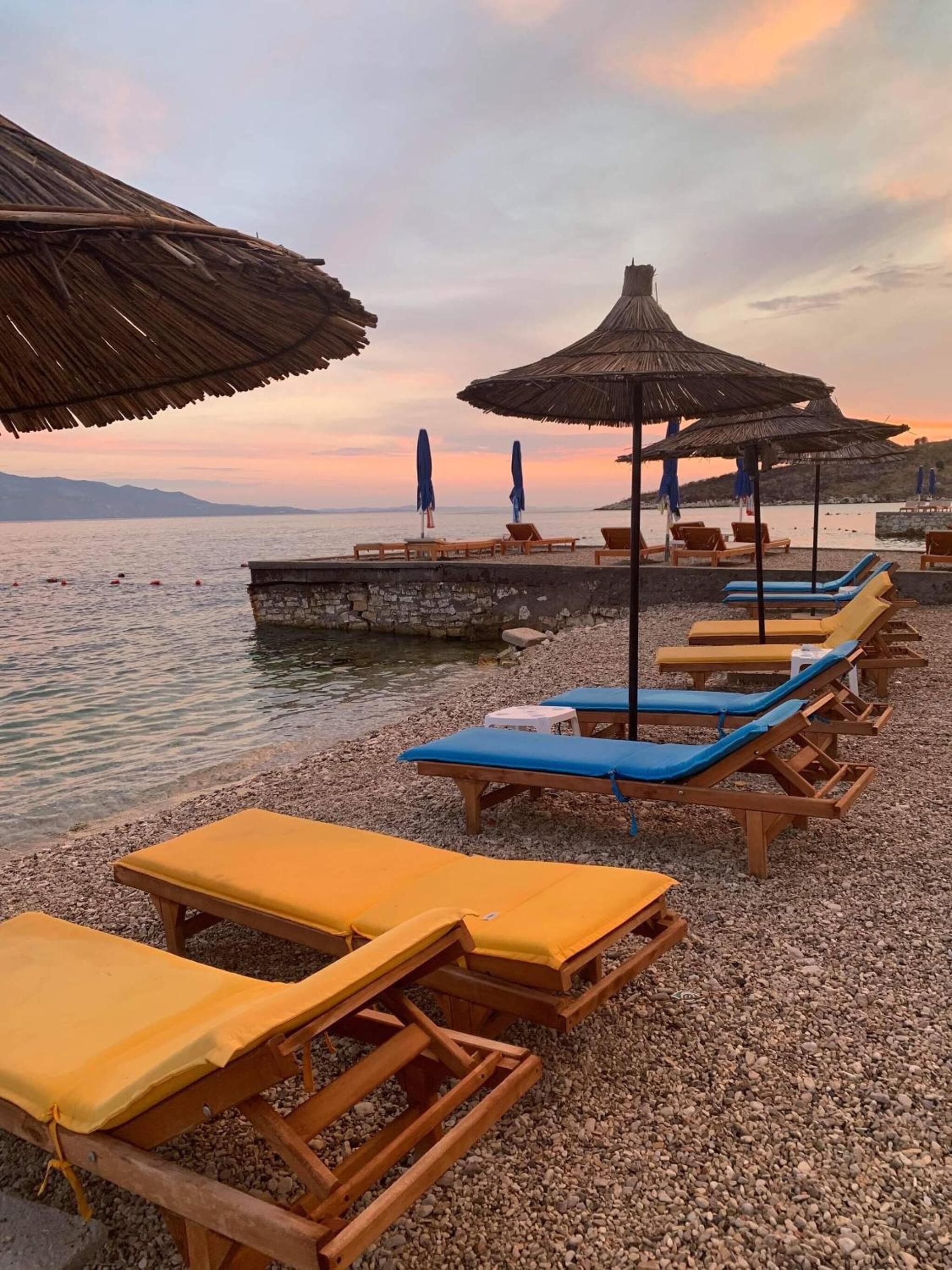A pebbled beach with wooden lounge chairs and thatched umbrellas, overlooking calm waters.