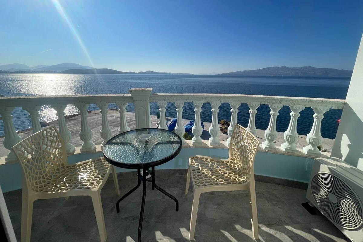 A balcony with a table and two chairs, overlooking a body of water and distant mountains.