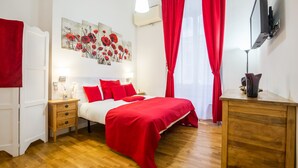 A bedroom with a wooden floor, a bed with red bedding, a nightstand, a dresser, a painting on the wall, and a TV mounted on the wall.