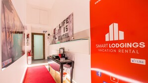 A modern hotel lobby with a red carpet, a glass partition, and a 'Life is Beautiful' sign. There's a desk with a chair and a small table with a plant.
