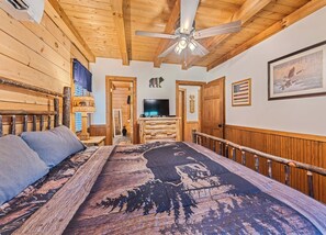 A room with a bed, a television, wooden walls, a ceiling fan, and a door leading to another room. - 2BD 2BA Cozy Log Cabin, Easy Paved Access, View, Gas Log, Hot Tub, Wifi Pond (Waynesville)
