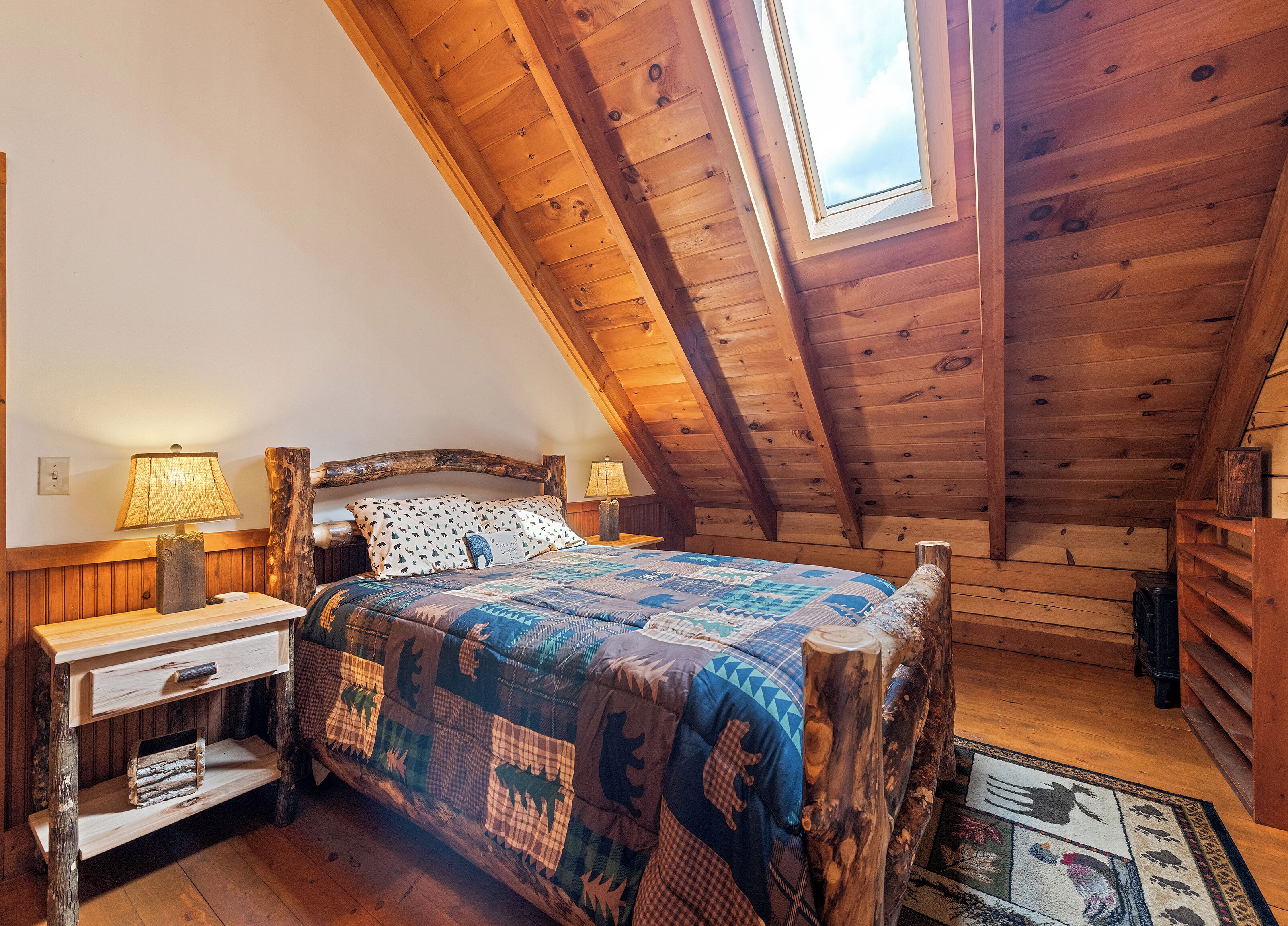 A wooden loft bedroom with a sloped ceiling, a bed with a patterned quilt, two bedside lamps, and a rustic wooden nightstand.