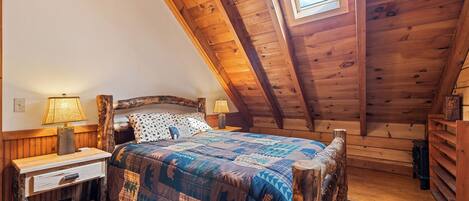 A wooden loft bedroom with a sloped ceiling, a bed with a patterned quilt, two bedside lamps, and a rustic wooden nightstand.