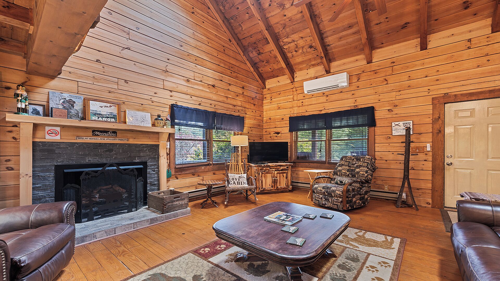A cozy cabin interior with a fireplace, leather sofas, a television, and a rustic wooden ceiling.