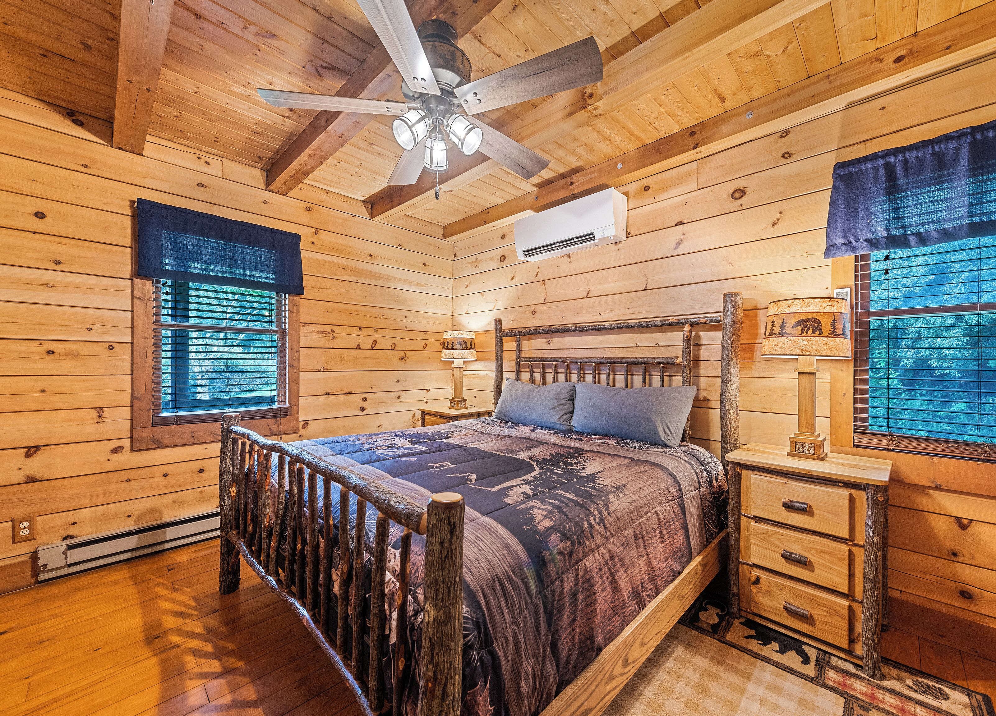 Un dormitorio de cabaña de madera con una cama, una mesita de noche, una lámpara, un ventilador de techo y una ventana con persianas.