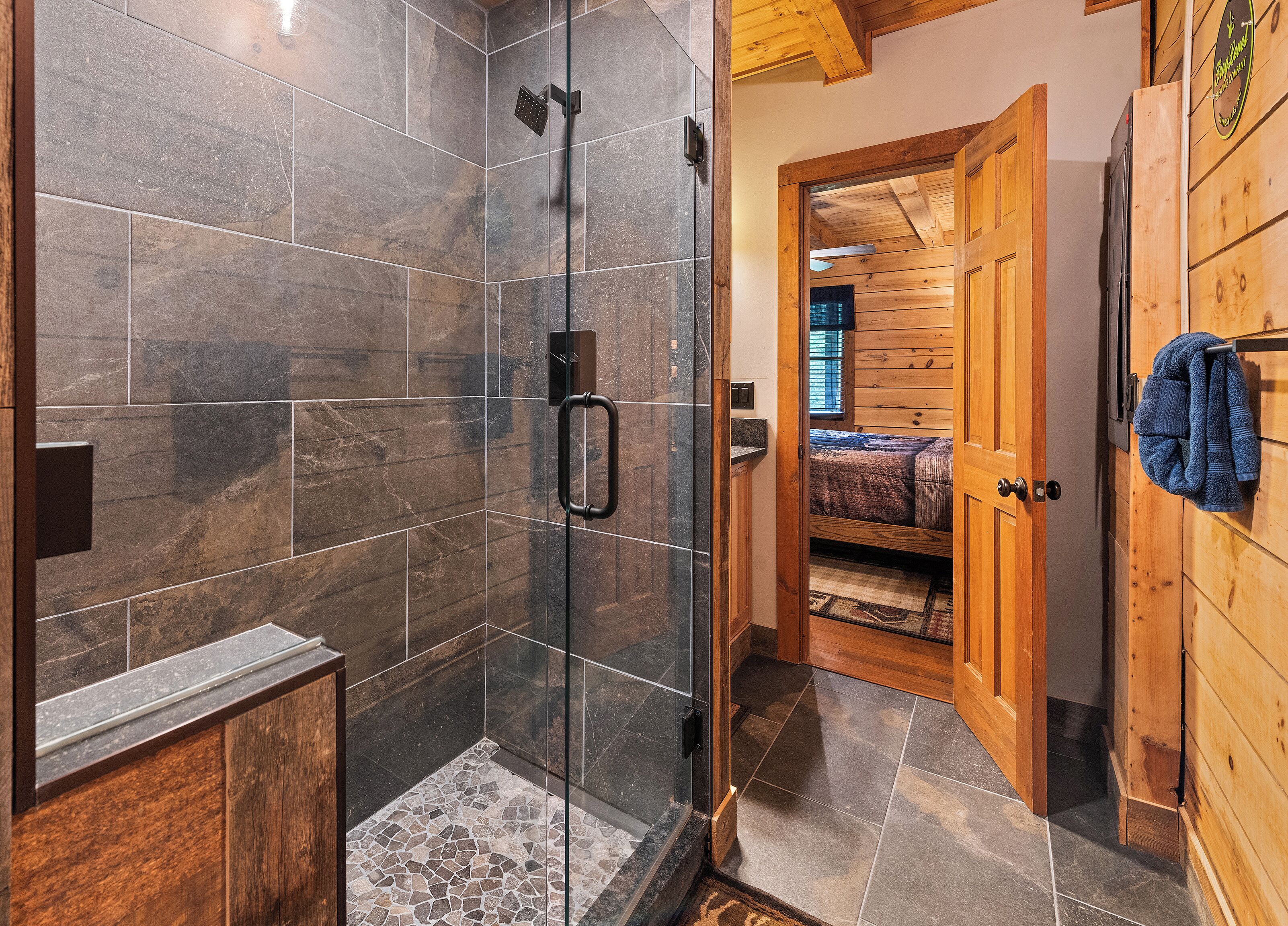 A modern bathroom with a glass-enclosed shower, a wooden door leading to a bedroom, and a towel hanging on the wall.