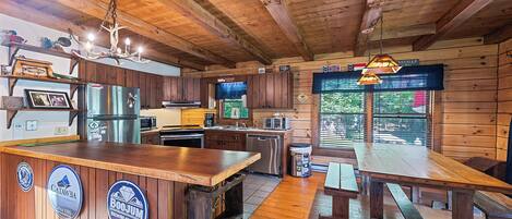 A rustic cabin interior with wooden walls, a wooden ceiling, a kitchen with modern appliances, a bar area, and a dining table.
