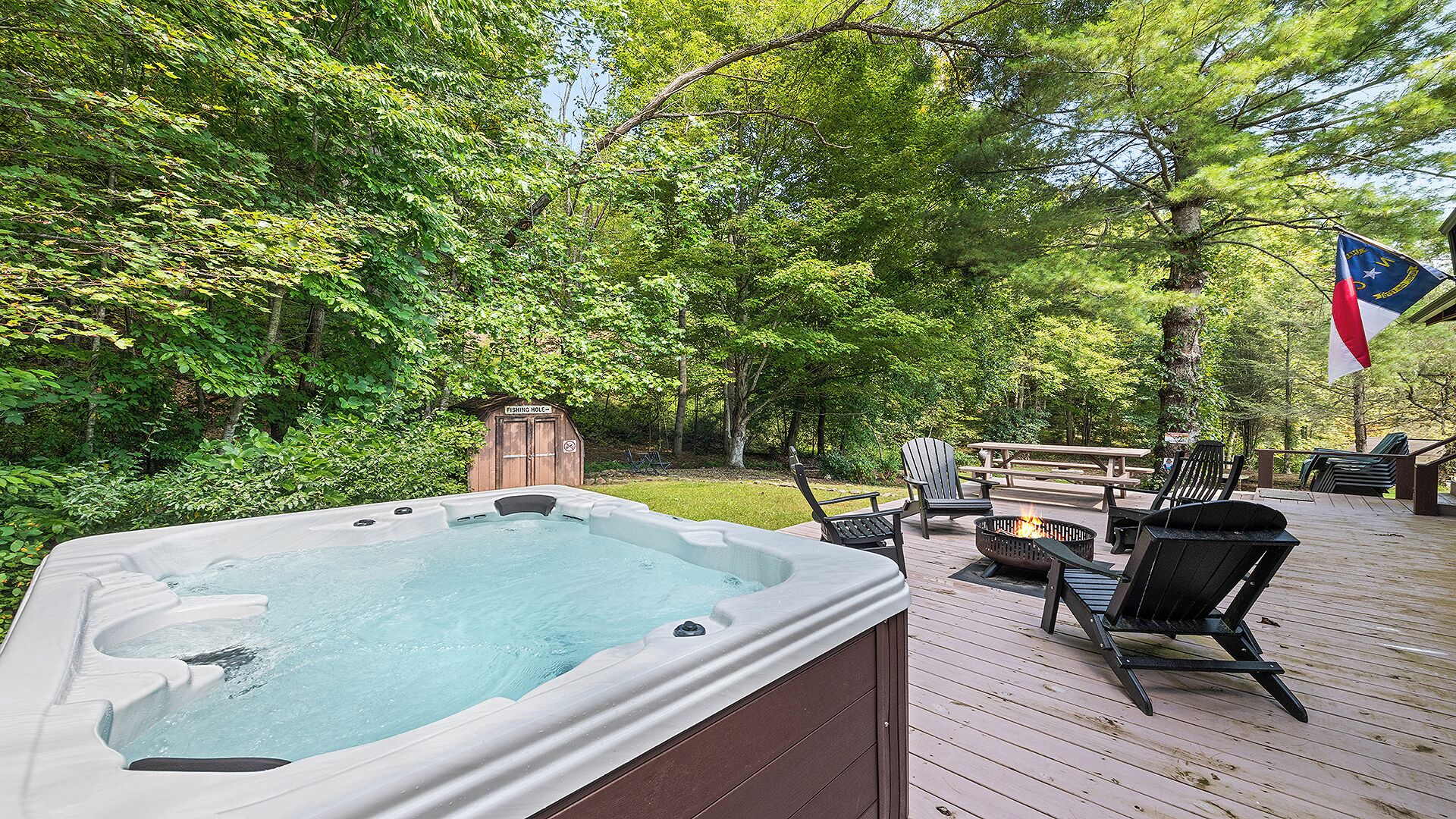 A hot tub on a wooden deck surrounded by lush greenery, with outdoor seating and a fire pit.