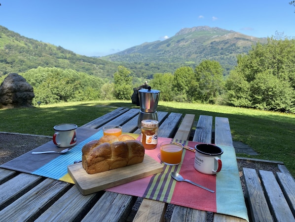 Un montaje para picnic con hogaza de pan, una termo, una taza y un tarro de miel sobre una mesa de madera, con una vista panorámica de montañas al fondo.