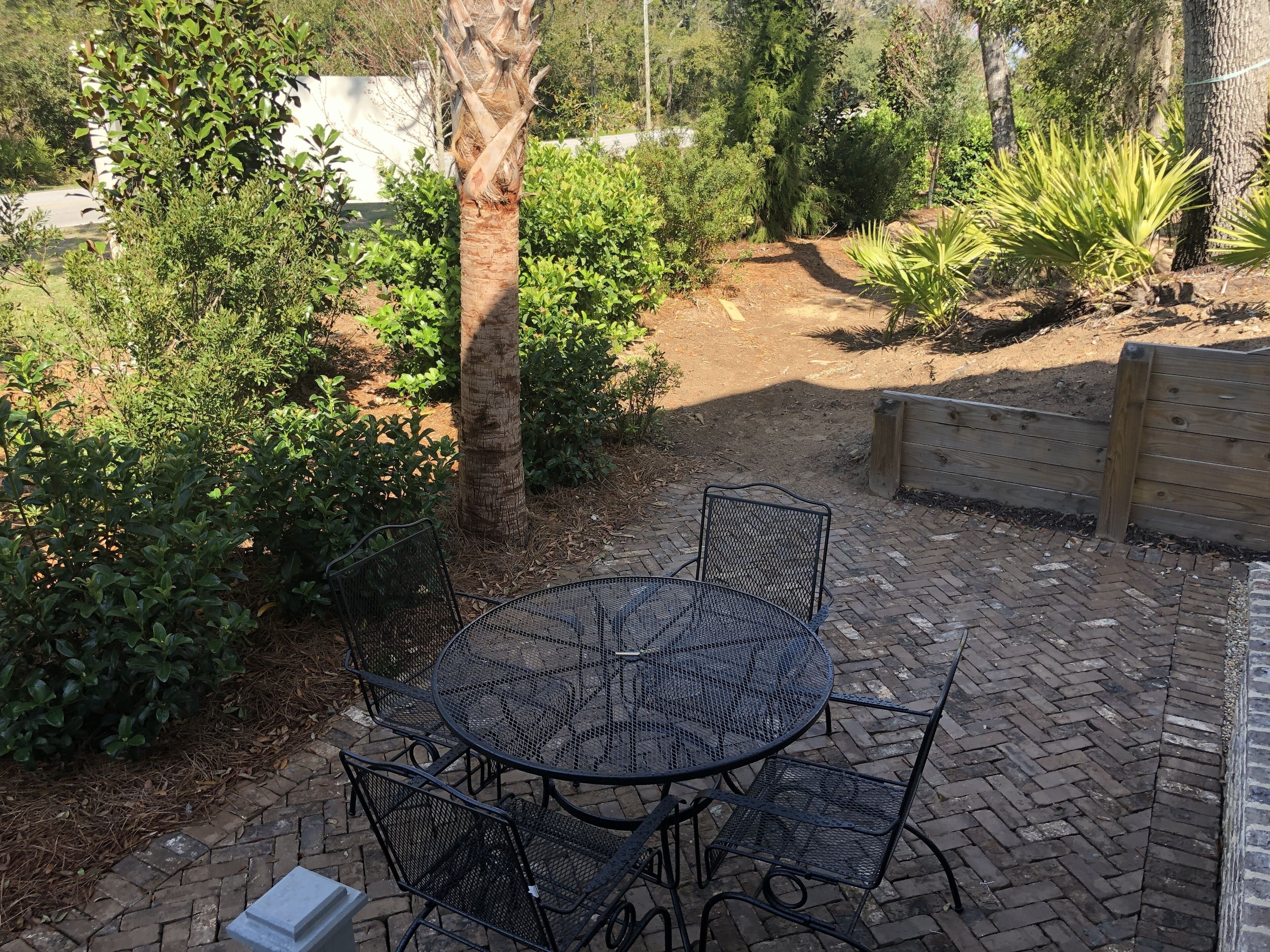 A patio with a round metal table and four chairs, surrounded by a brick pathway and various plants.