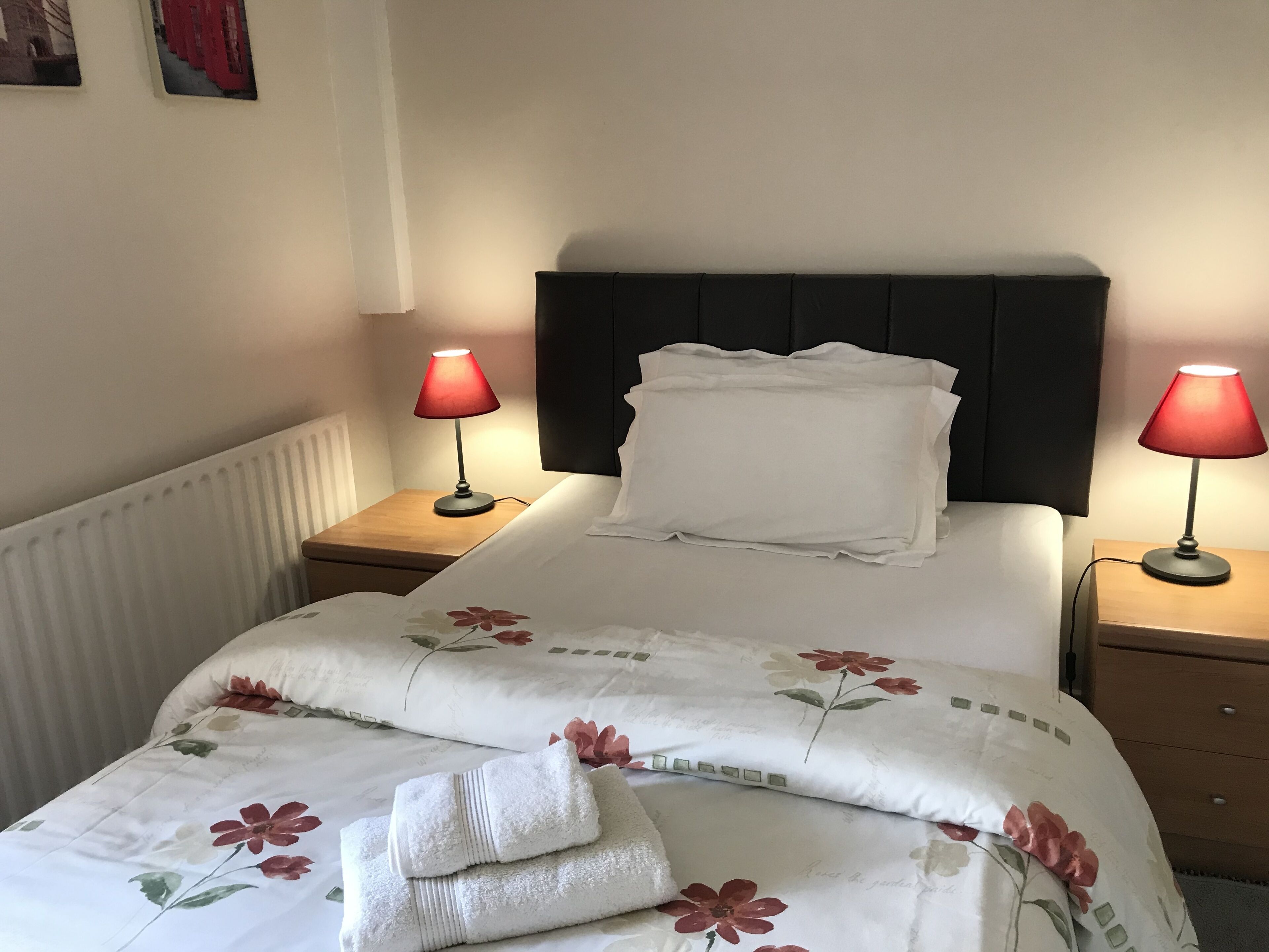 A neatly made bed with floral bedding, two bedside lamps, and a radiator.