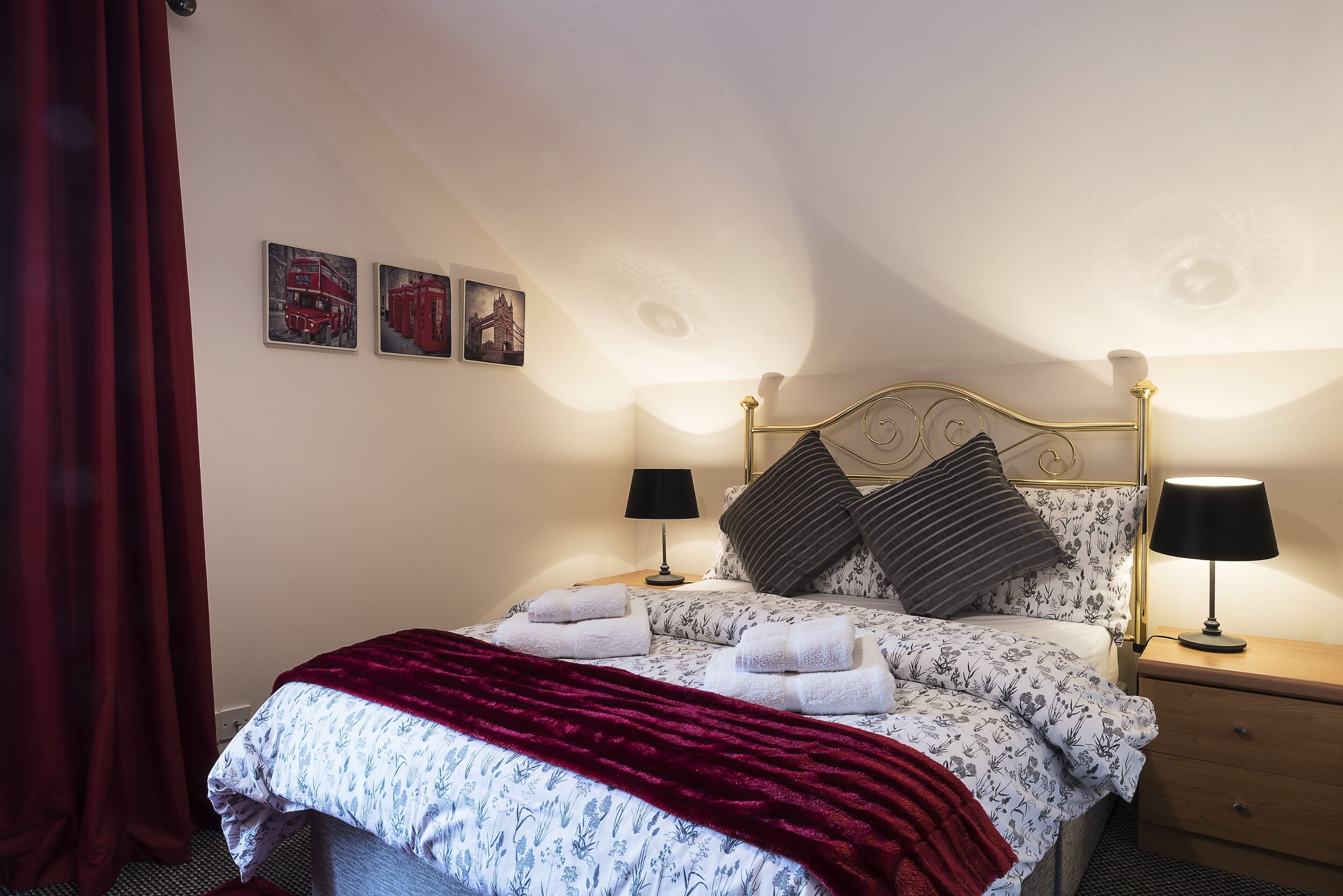A neatly made bed with a floral duvet, two pillows, and a red throw blanket. There are two bedside tables with lamps, and a red curtain is partially visible.