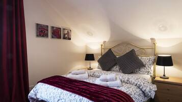 A neatly made bed with a floral duvet, two pillows, and a red throw blanket. There are two bedside tables with lamps, and a red curtain is partially visible.