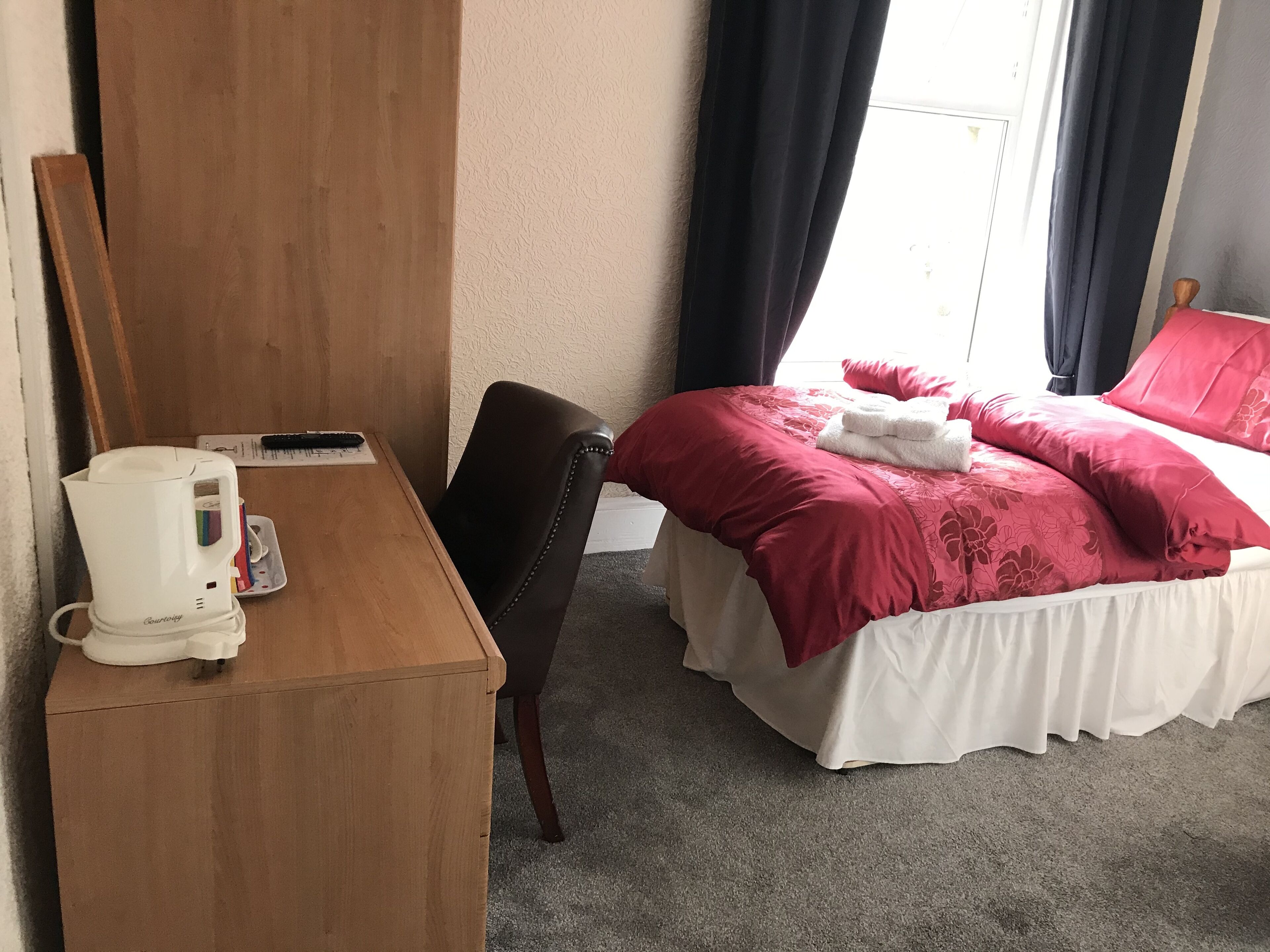 A hotel room with a wooden desk, a chair, a bed with red bedding, and a window with curtains.