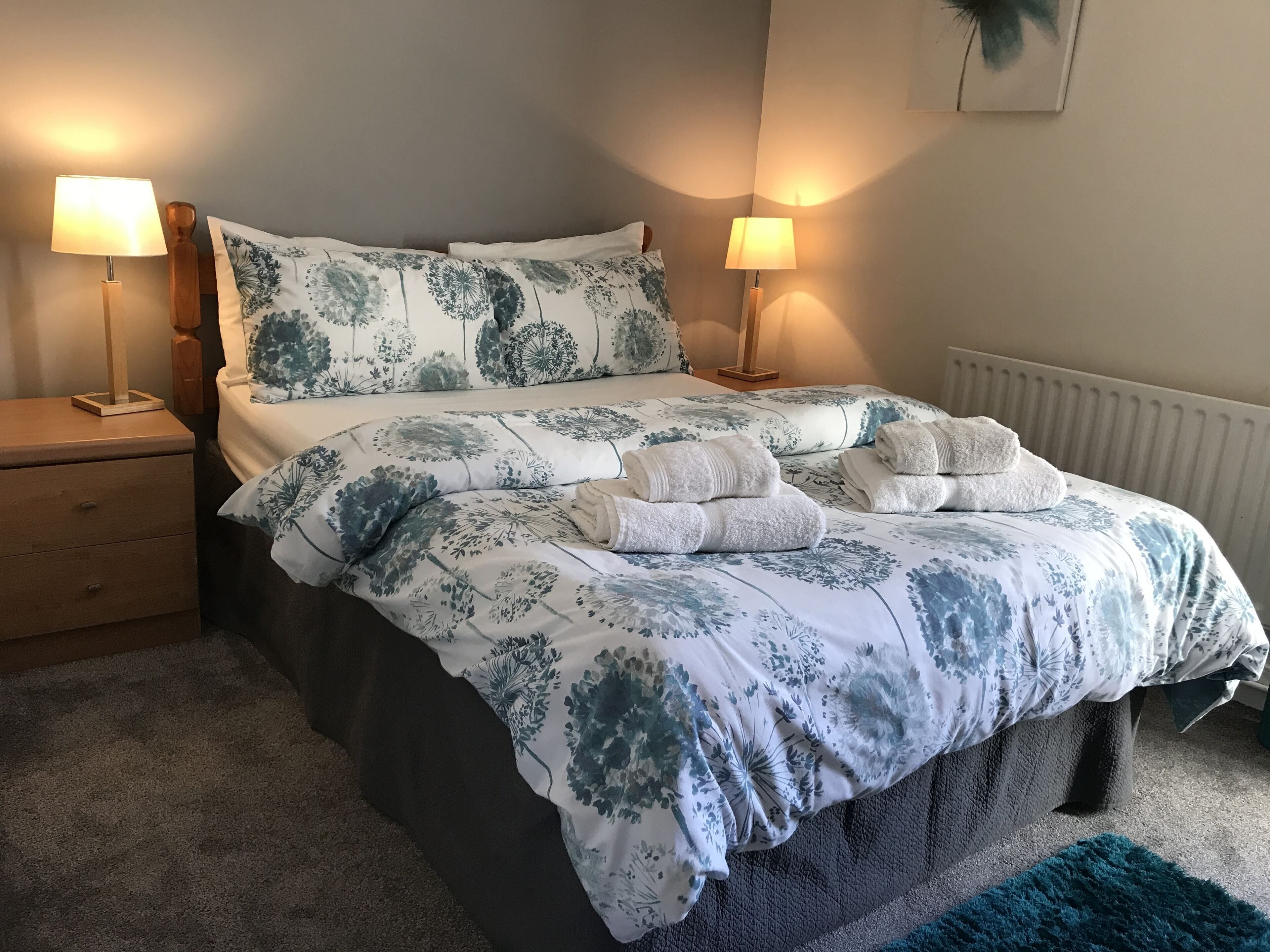A neatly made bed with floral bedding, two bedside lamps, a nightstand, and a radiator.