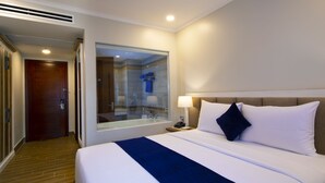 Habitación de hotel con una cama grande, una mesita de noche, una lámpara y vista al baño a través de una puerta de vidrio.