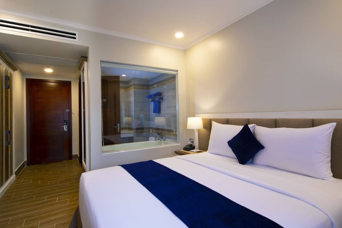 Une chambre d’hôtel avec un grand lit, une table de chevet, une lampe et une vue sur la salle de bain à travers une porte en verre.