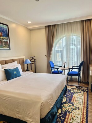 A hotel room with a large bed, a desk with two chairs, a painting on the wall, and a view through the window. - Sabina Hotel 2 (Ho Chi Minh City)