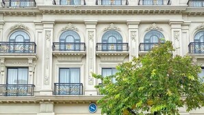 Fachada imponente de un hotel con detalles arquitectónicos ornamentales, varios balcones y una entrada central con asientos al aire libre.