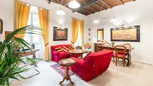 A living room with a red sofa, wooden dining table, and chairs. There is a potted plant, a small round table, and a painting on the wall.