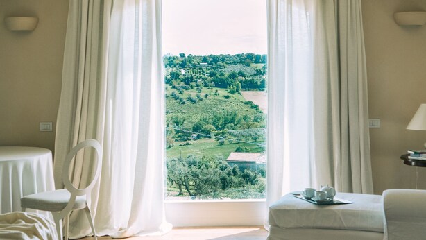 A room with a wooden ceiling, a large window with curtains, a bed, a chair, and a table with a tray. - Charm and hospitality at 500 meters from the sea. Pool surrounded by greenery (Civitanova marche)