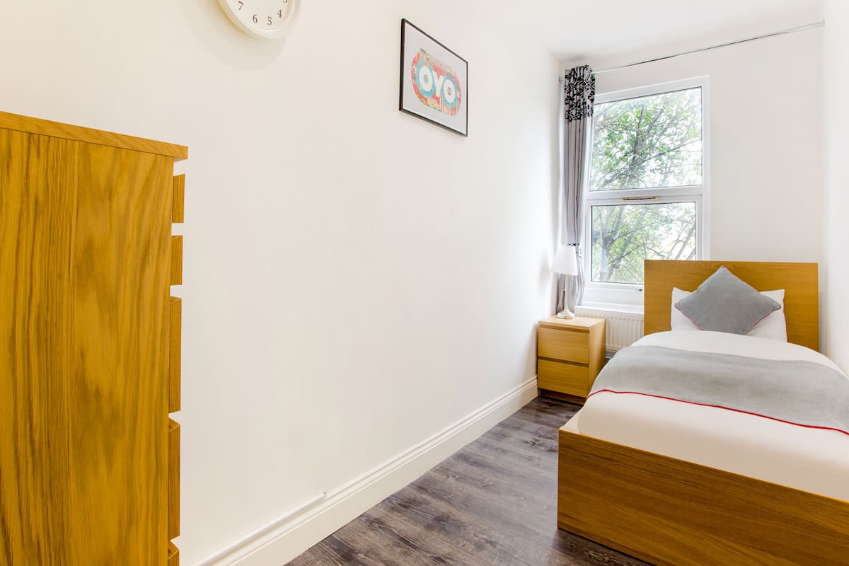 A bedroom with a wooden wardrobe, a bed with a headboard, a bedside table, a lamp, a clock, and a window with a view of trees.