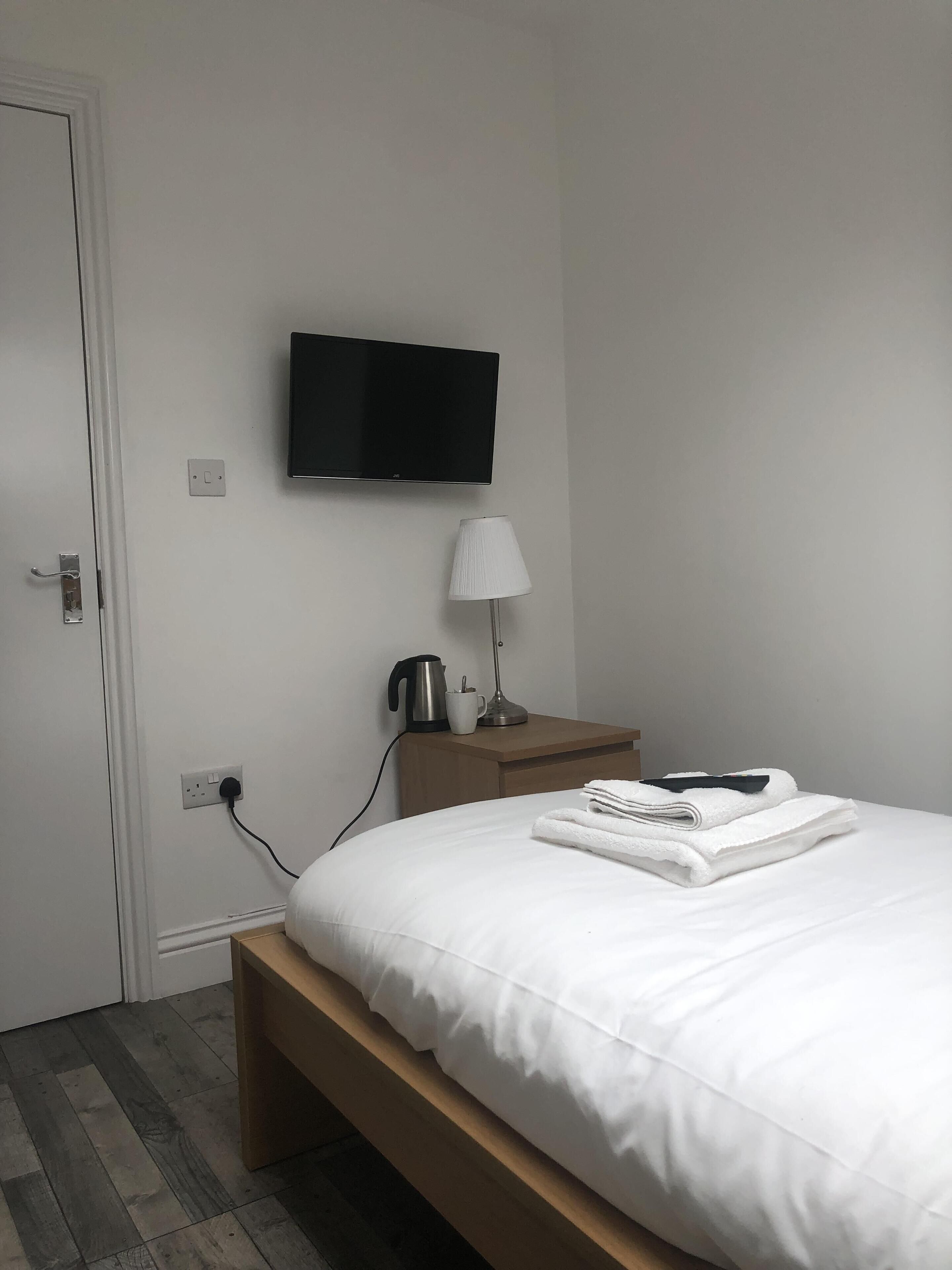A hotel room with a bed, a television mounted on the wall, a bedside table with a lamp, a kettle, and a cup.