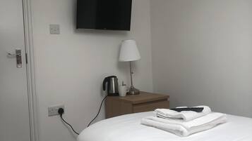 A hotel room with a bed, a television mounted on the wall, a bedside table with a lamp, a kettle, and a cup.