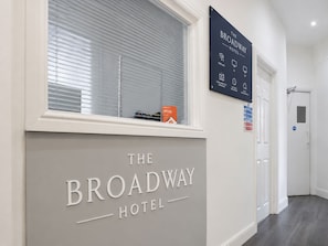 A hallway with a sign for The Broadway Hotel, a window with blinds, and a door. - The Broadway Hotel - London Heathrow (Southall)