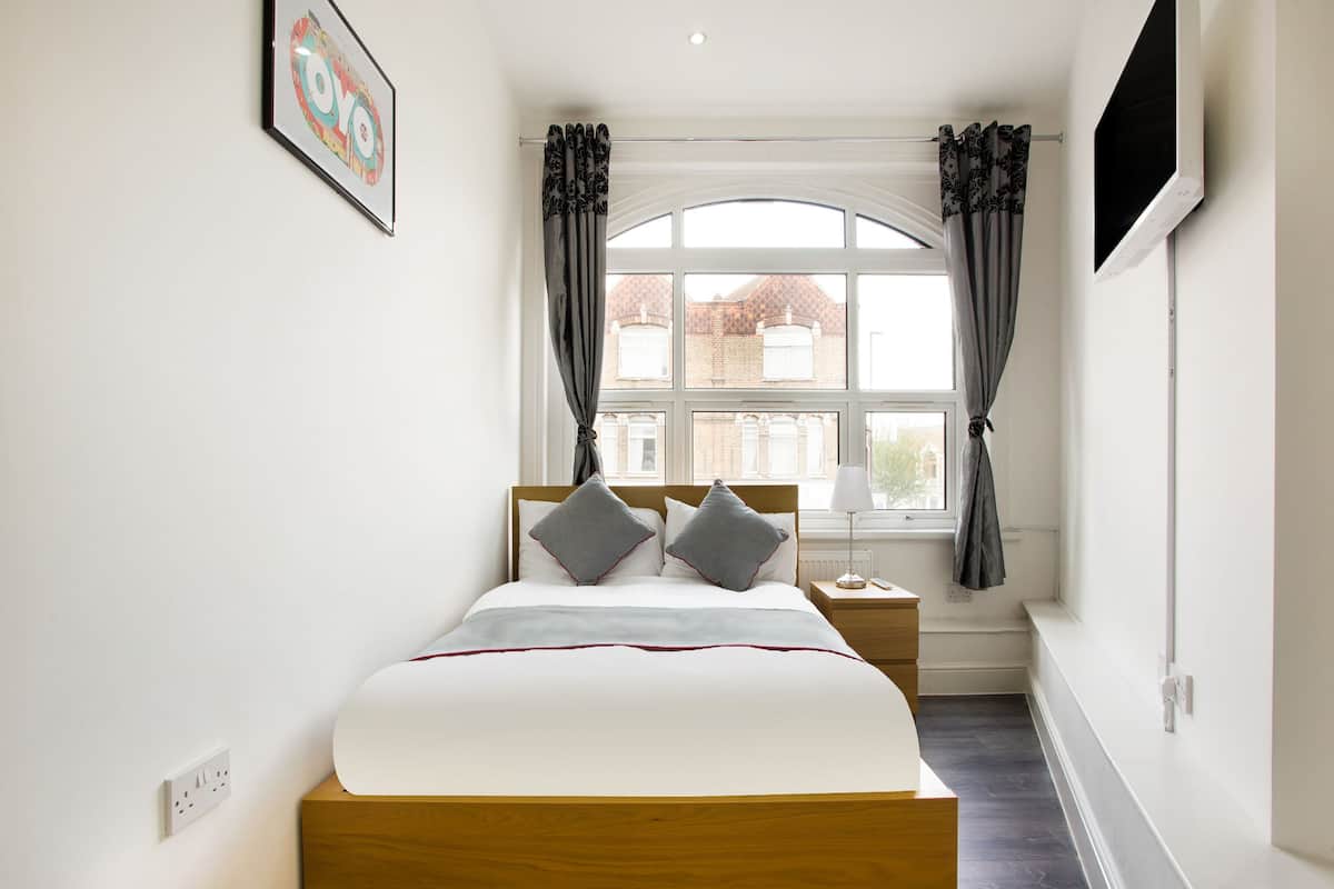 A bedroom with a bed, a bedside table, a lamp, a TV mounted on the wall, and a window with curtains.