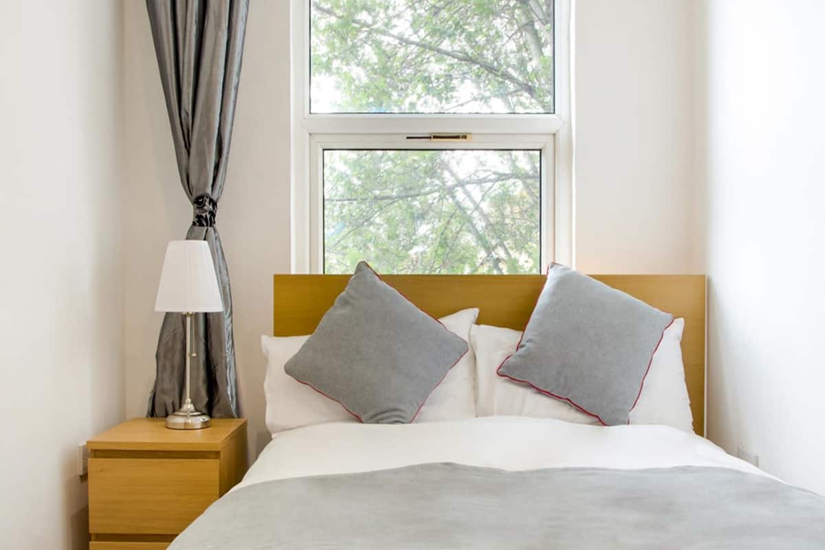 A neatly made bed with a grey and white bedding set, a wooden nightstand with a lamp, and a window with a view of trees.