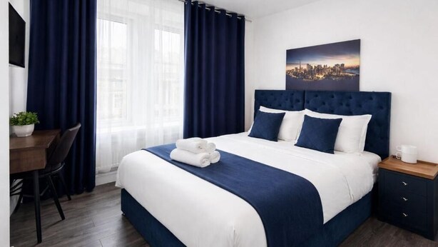 A hotel room with a large bed, a desk with a chair, a small table, and a nightstand. - The Broadway Hotel - London Heathrow (Southall)
