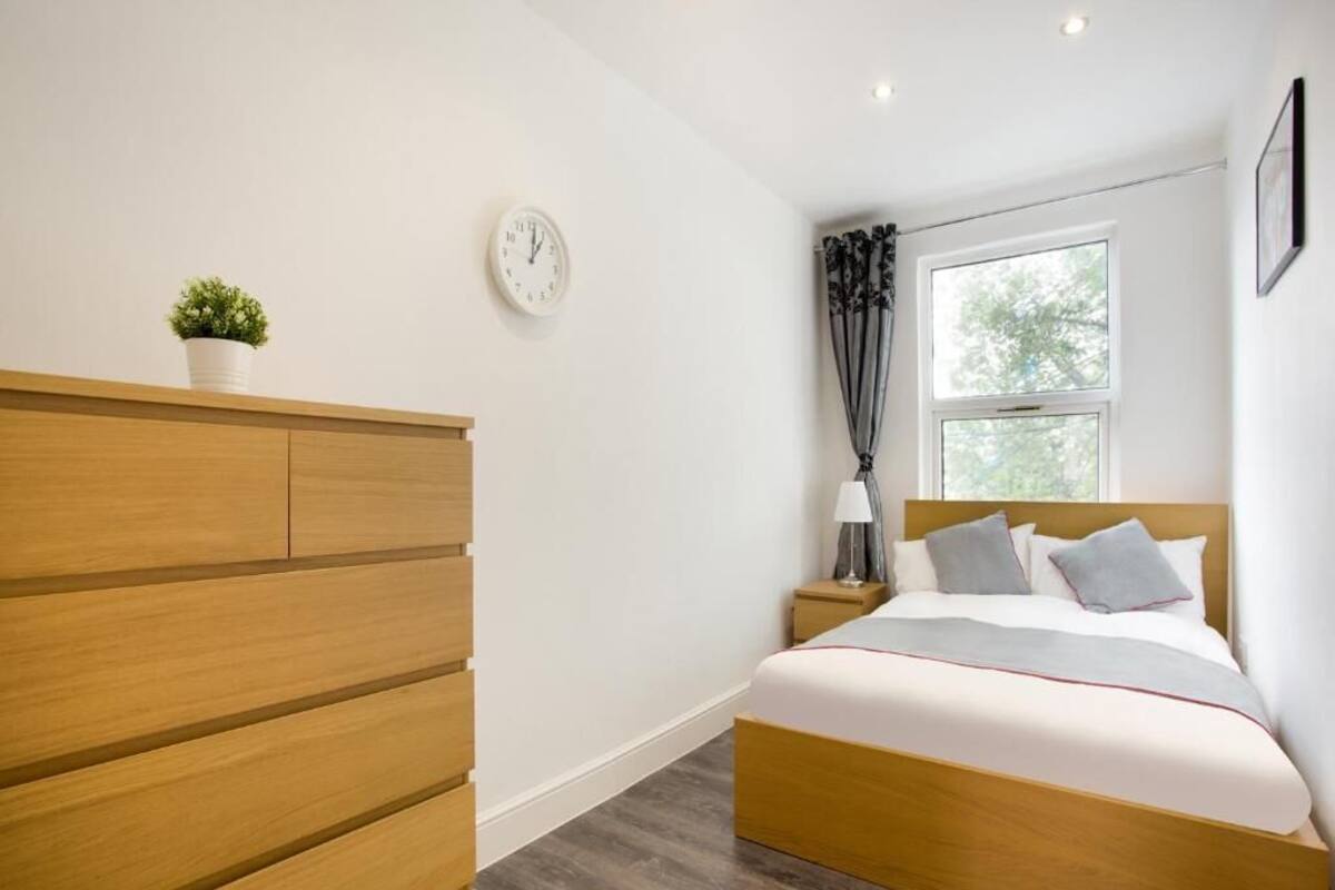 A bedroom with a wooden bed, a bedside table with a lamp, a clock on the wall, and a window with curtains.