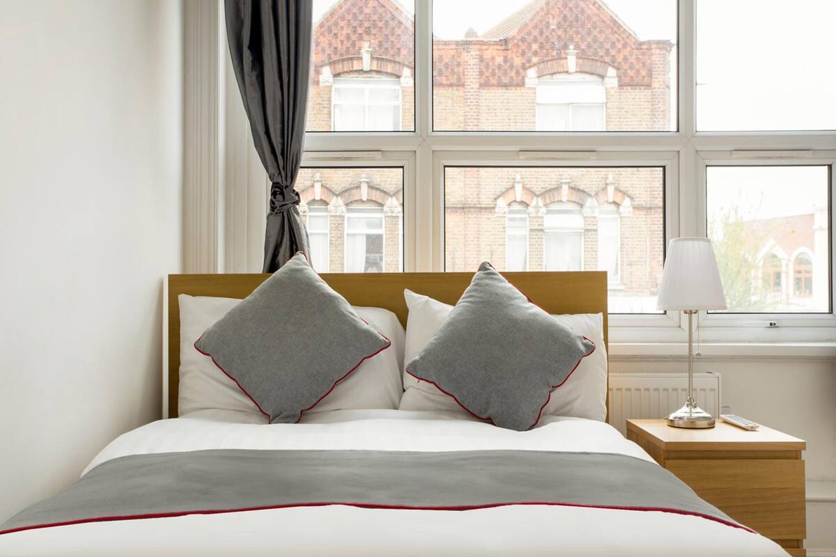 A neatly made bed with a grey and white color scheme, a wooden headboard, and a view of a brick building through a window.