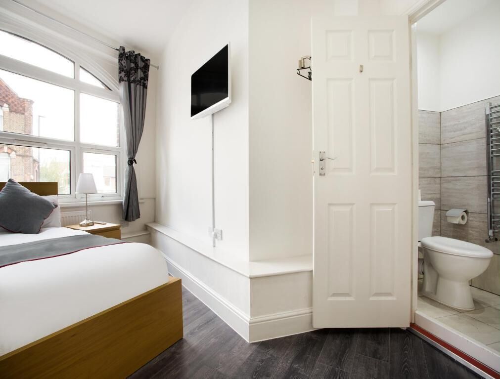A bedroom with a bed, a TV mounted on the wall, a door leading to a bathroom, and a window with curtains.