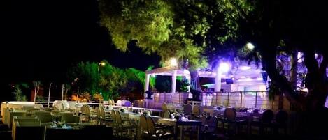 Un área de comedor al aire libre con mesas y sillas dispuestas bajo una sección cubierta, rodeada de árboles e iluminación ambiental.