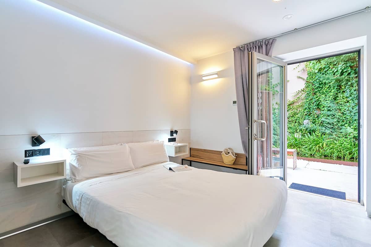 A modern hotel room with a large bed, bedside tables, a nightstand, and a view of a lush garden through a sliding glass door.