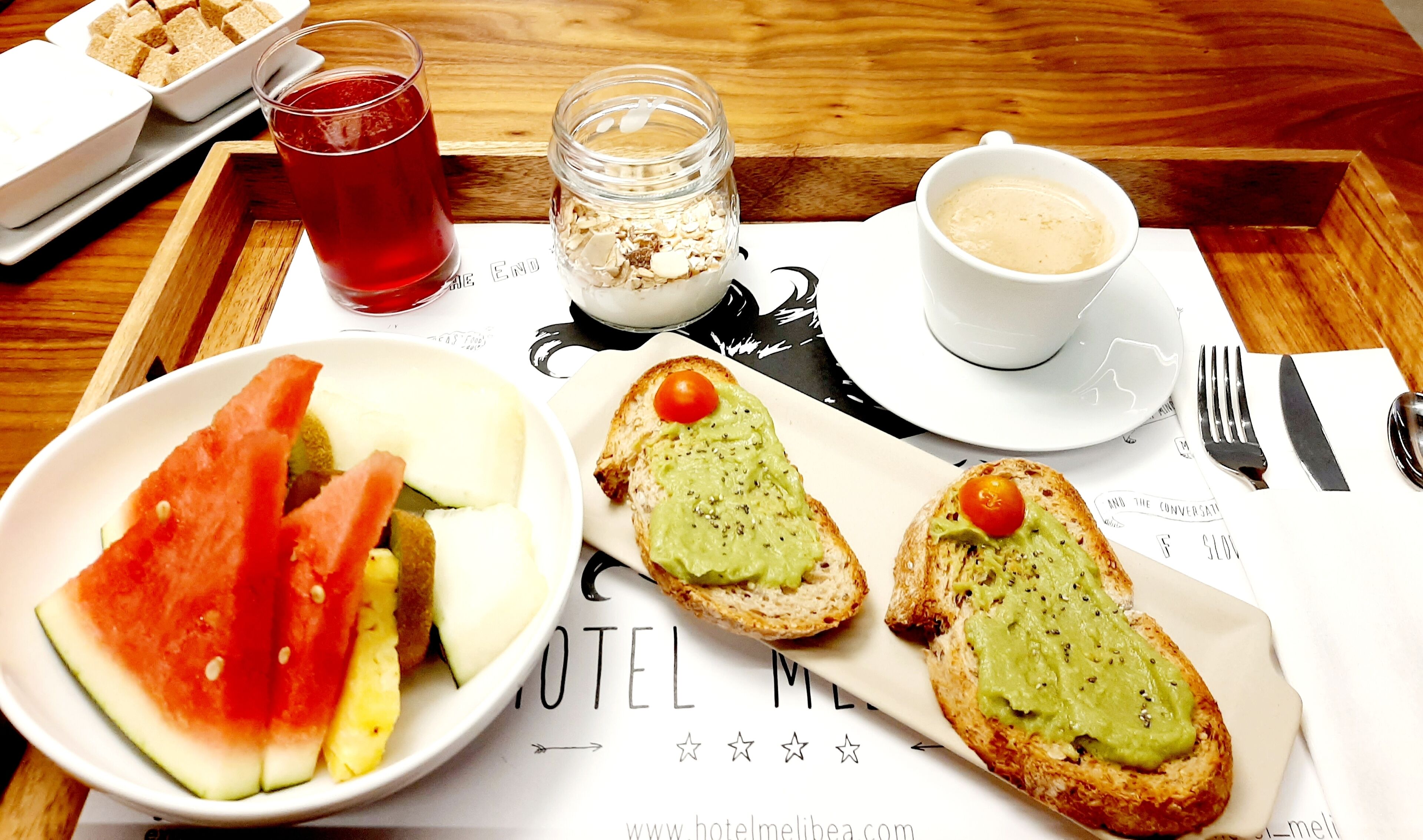 A breakfast tray with toast, avocado, tomato, and watermelon, a cup of coffee, and a glass of red beverage.