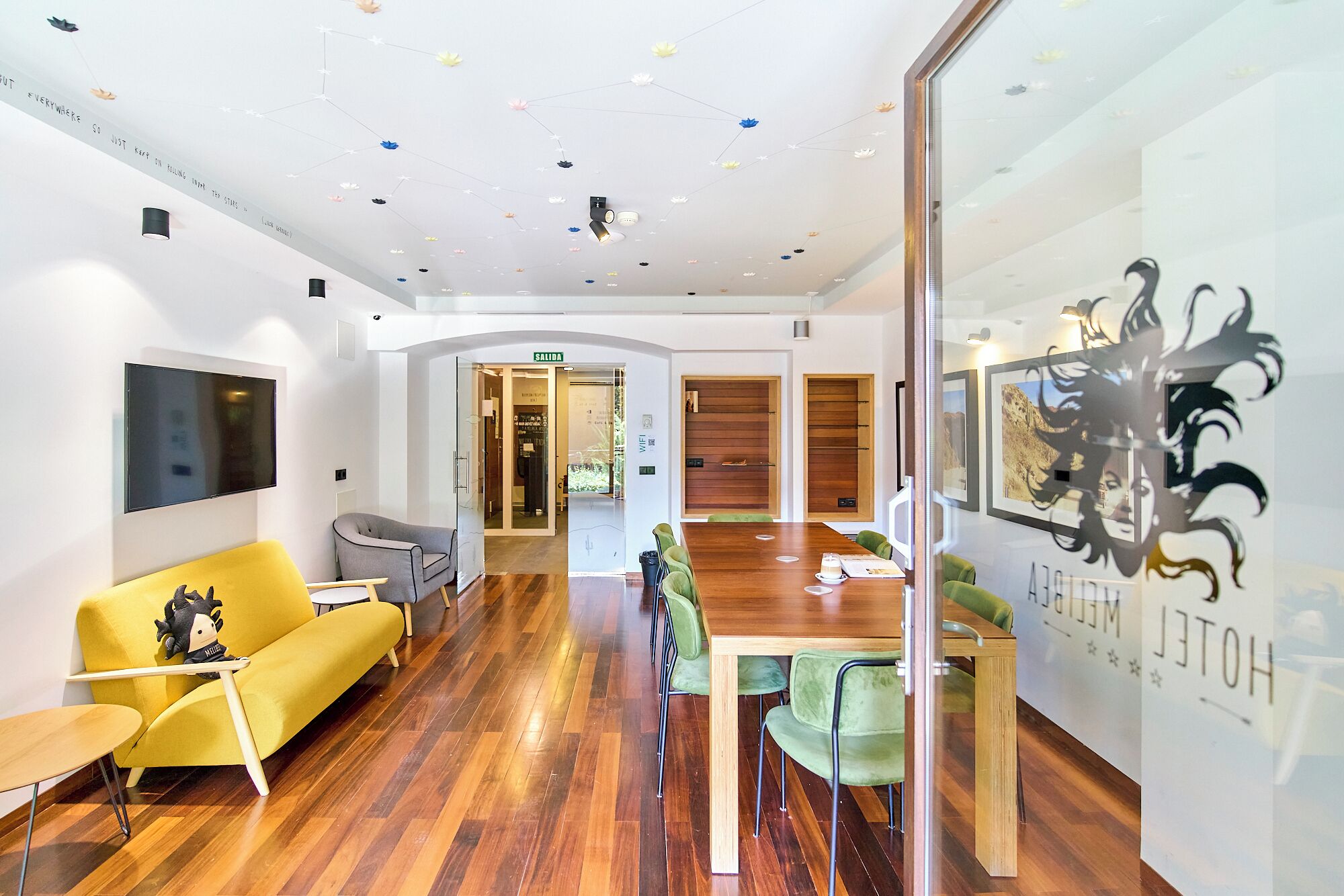 Un interior moderno con piso de madera, un sofá amarillo, una mesa de madera y una pared de vidrio con un logotipo.