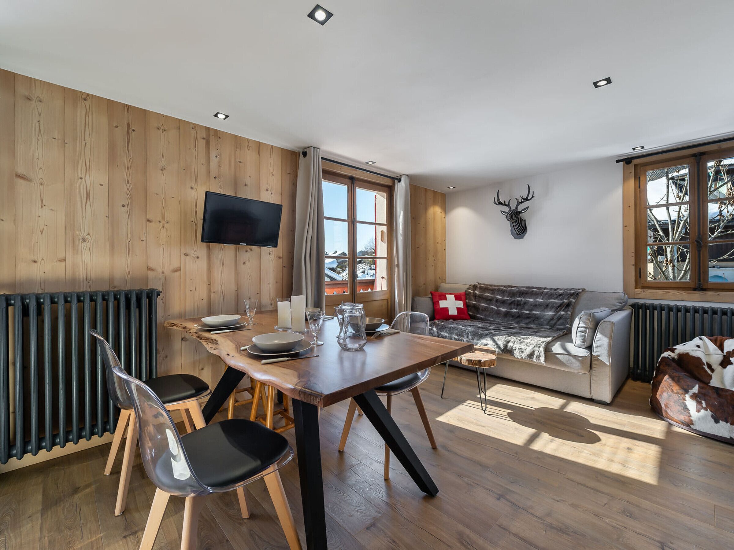 A modern living room with a wooden floor, a dining table with chairs, a sofa, and a mounted TV.