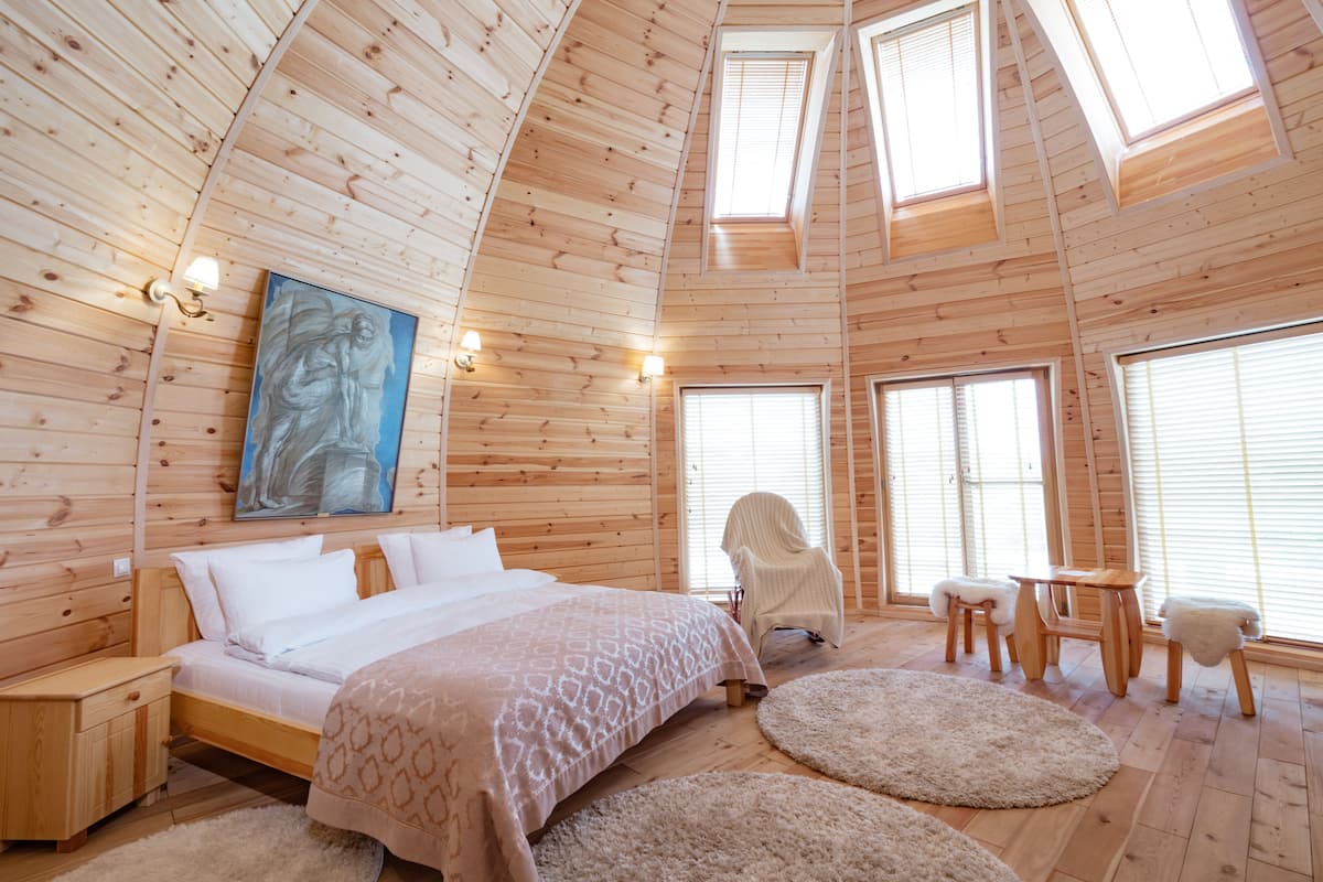 A wooden cabin interior with a bed, a chair, a table, and a painting on the wall.