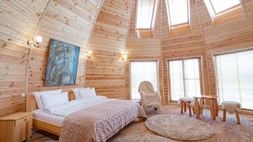 A wooden cabin interior with a bed, a chair, a table, and a painting on the wall.