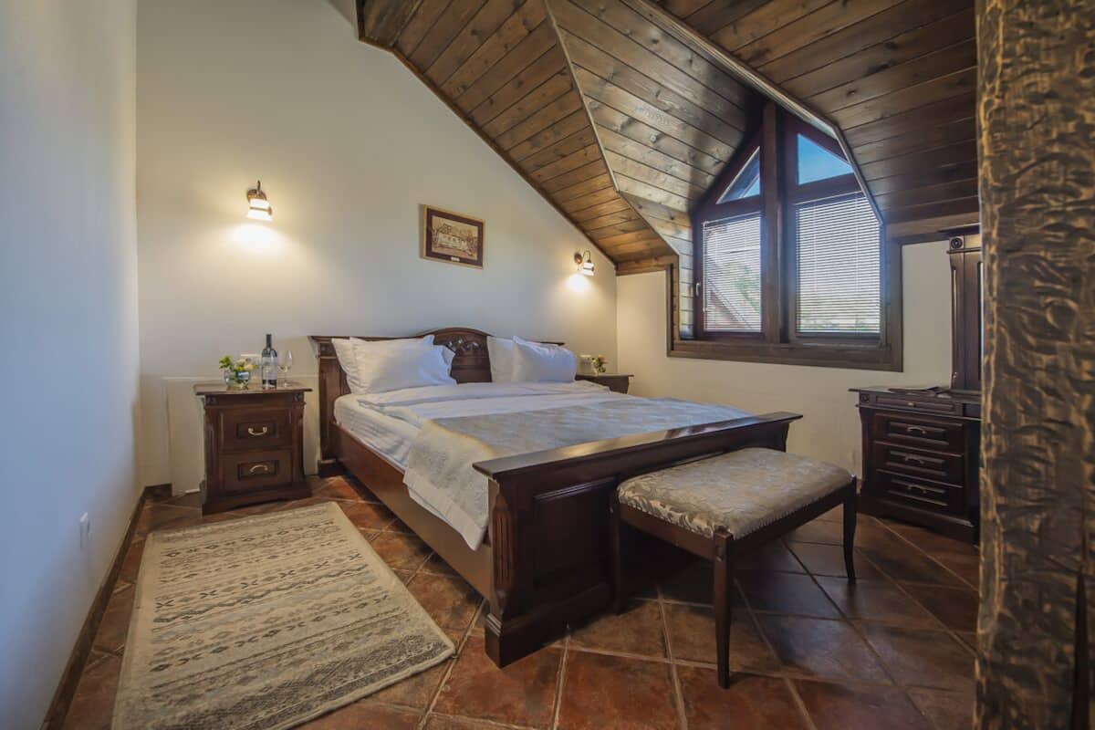 A bedroom with a wooden ceiling, a large bed, a bedside table, a bench, and a window with blinds.