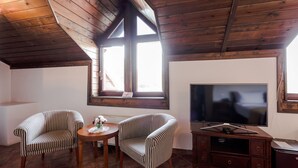 Una habitación acogedora con vigas de madera en el techo, un ventanal grande, dos sillones a rayas, una mesita redonda y un televisor sobre un mueble de madera.