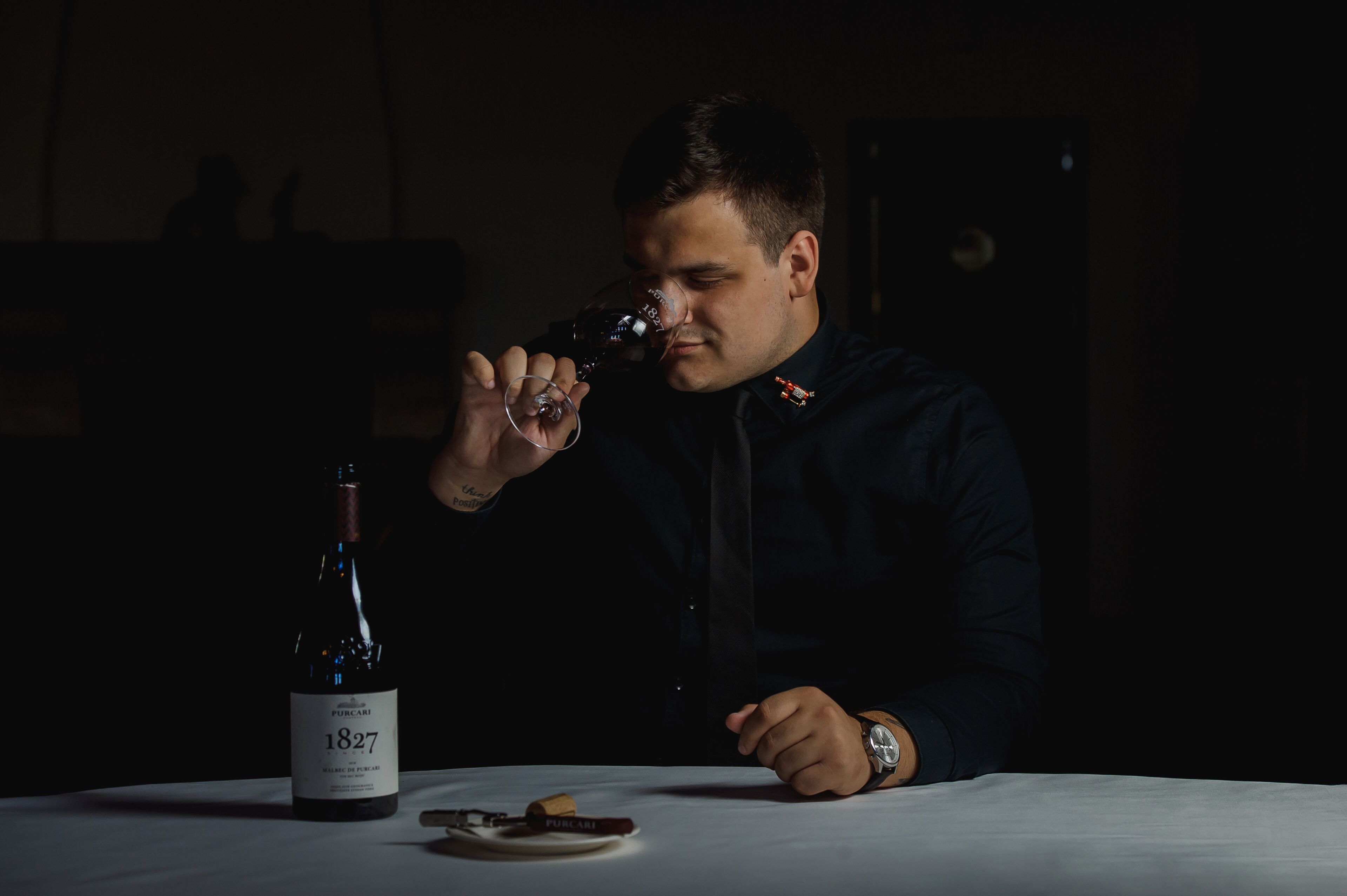 A person seated at a table with a bottle of wine labeled "1827" and a corkscrew.
