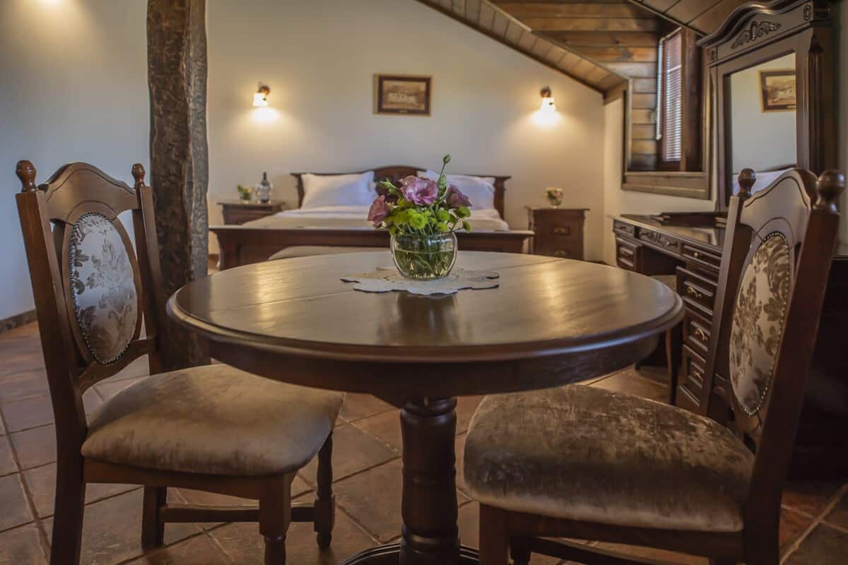 A dining area with a round table, two chairs, and a vase with flowers. There is a bed with a wooden headboard in the background.